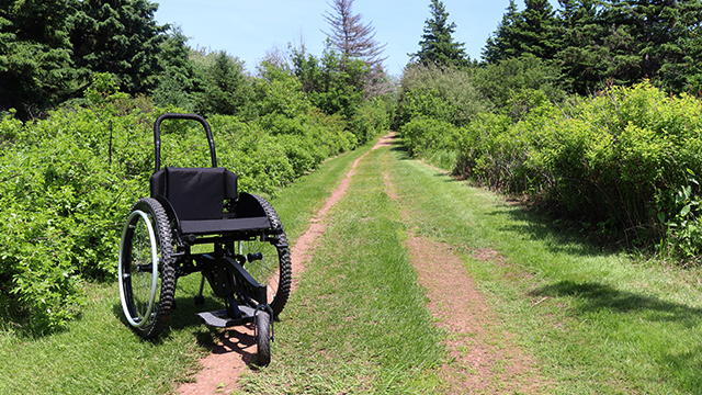 A modified wheelchair with one smaller wheel in front that pivots, sits a besides a trail surround by lush green trees and grass on a sunny day.