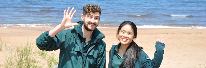 Deux étudiants de Parcs Canada en uniforme se tiennent debout et saluent à l'entrée de la plage Dalvay, dans le parc national de l'Île-du-Prince-Édouard. 