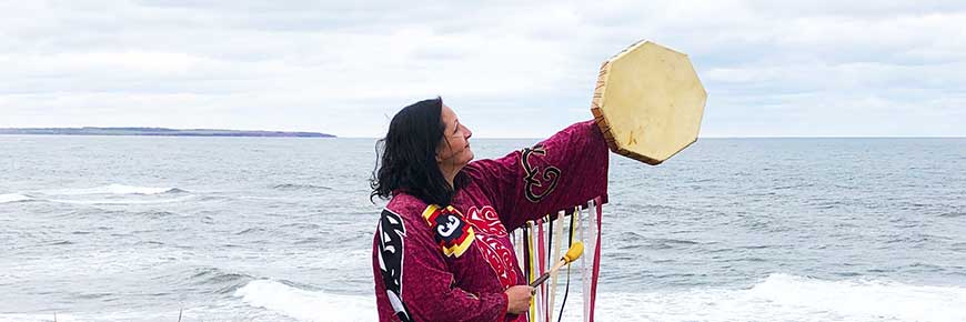 Julie Pellissier-Lush, a Mi'kmaq Knowledge Keeper, stands in full regalia holding her hand drum up, on a cliff in front of the ocean. 