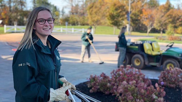 Un jeune vêtu d'un uniforme de Parcs Canada se tient debout près d'un bac rempli de plantes.  À l'arrière-plan, d'autres personnes travaillent par une journée ensoleillée. 
