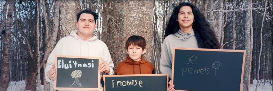 Trois jeunes hommes brandissent des pancartes disant : Elui'tmasi, I Promise, et Je Promets, tout en se tenant dans une forêt d'hiver avec beaucoup de neige et d'arbres derrière eux. 