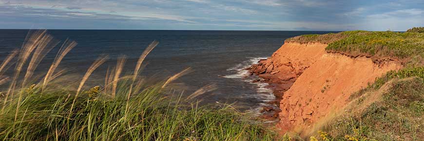 A red cliff at Oceanview in Prince Edward Island National Park, with a green grassy cliff in the foreground and a calm blue ocean and skyline in the background. 