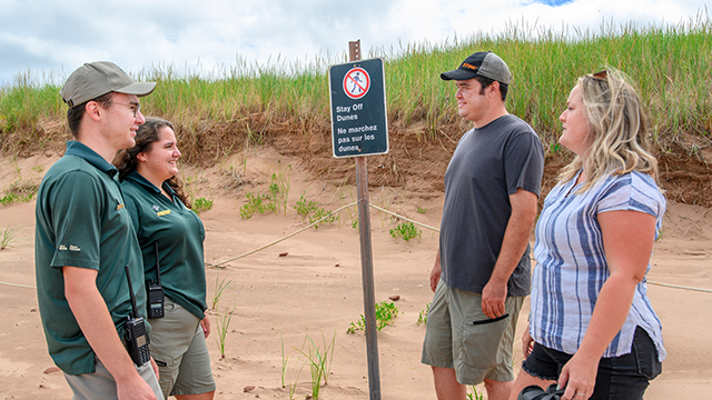 Two Parks Canada staff talk to two visitors beside a sign next to sand dunes on a sunny day.