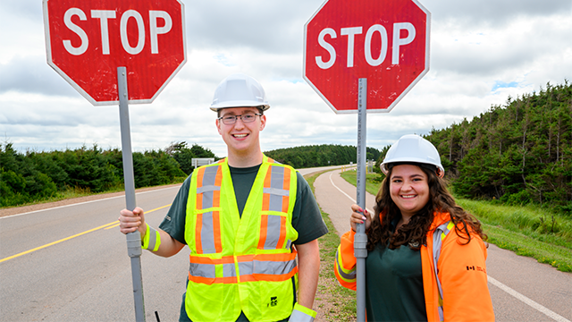 Two Parks Canada staff hold stop signs along the side of the road on a cloudy day.