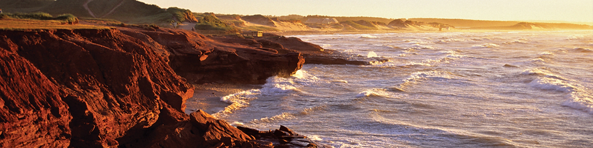 Tall red cliffs jut out into the ocean.  The sun sets in the distance.
