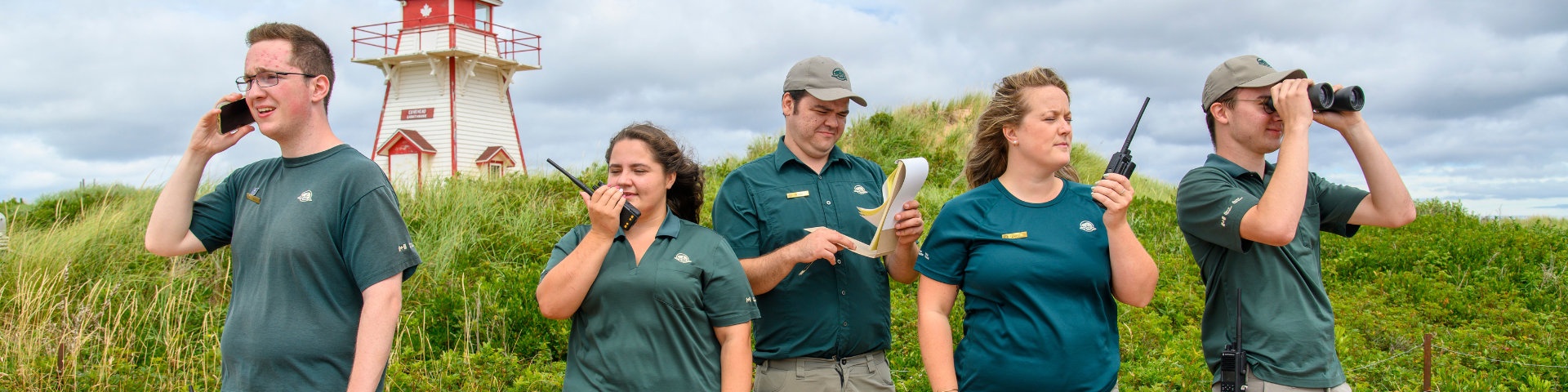 Five Parks Canada staff members wearing green shirts, stand in front of a dune with a red and white light house in the background.