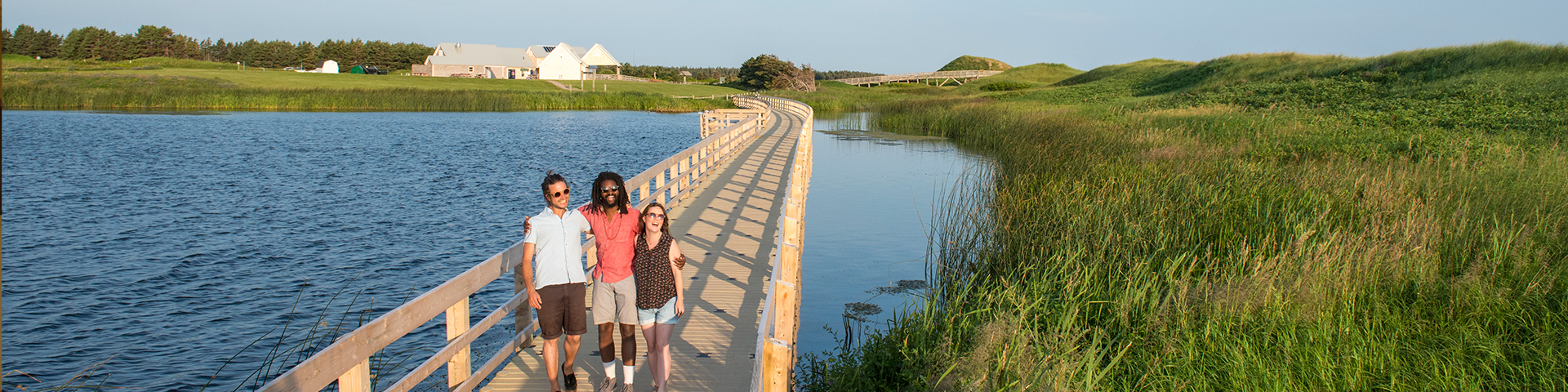 Visitors walk across a floating boardwalk on Cavendish Duneslands Trail with the Cavendish Beach facilities in the background on a summer day in Prince Edward Island National Park.