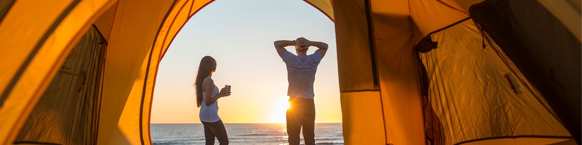 Two people stand outside a yellow tent in front of a body of water on a sunny day. 