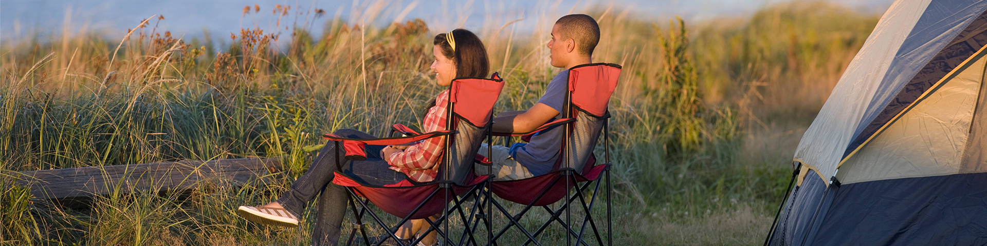 Two adults relaxing on camp chairs next to a tent overlooking the ocean.
