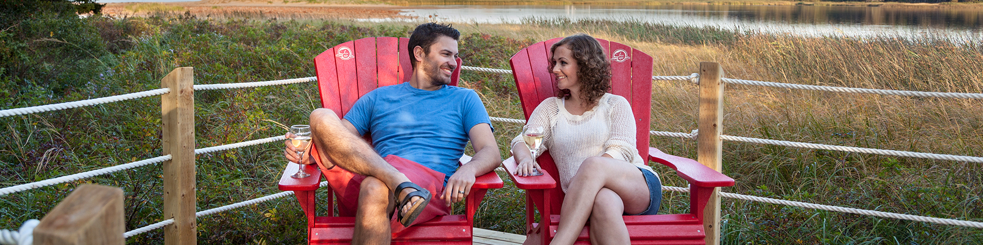 A couple relaxes in the red chairs at the lookout at Cavendish Campground on a summer evening with sand dunes in the background. Prince Edward Island National Park