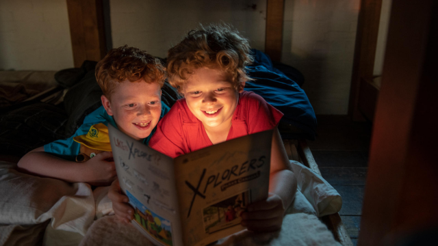 Two kids read a book together with a flashlight.
