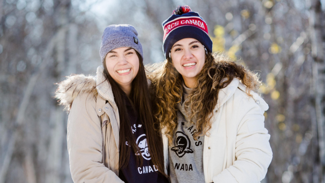 Two teens stand side by side wearing winter parkas and Parks Canada merchandise surrounded by winter trees. 