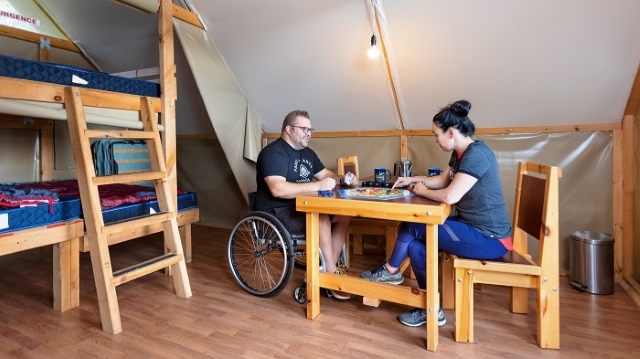 A person in a wheelchair and his partner are seated at a table in an otentik tent. 
