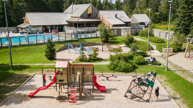 Des enfants s'amusent dans une aire de jeu devant un bâtiment de bois et une piscine. 