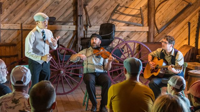 Three costumed entertainers make music in a barn in front of an audience. 