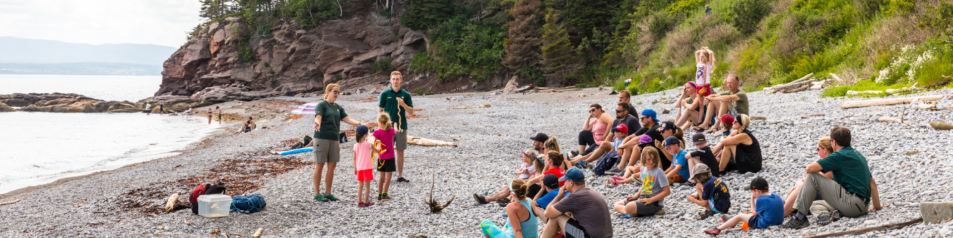 A group of visitors is participating in an activity with two Parks Canada employees on a pebble beach. 