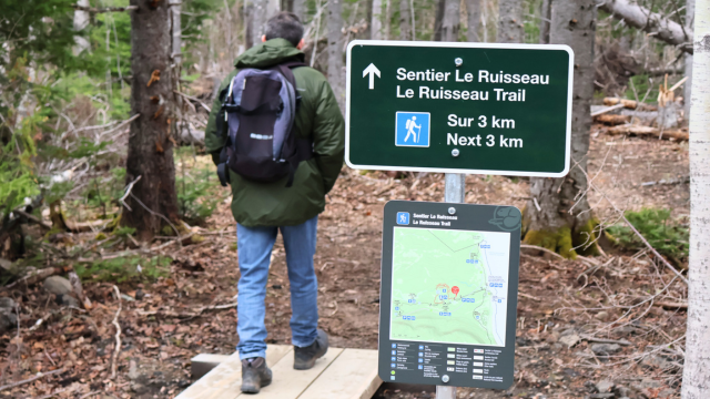 Un randonneur marche sur un ponceau et passe près d'un panneau où l'on voit le nom et la carte du sentier