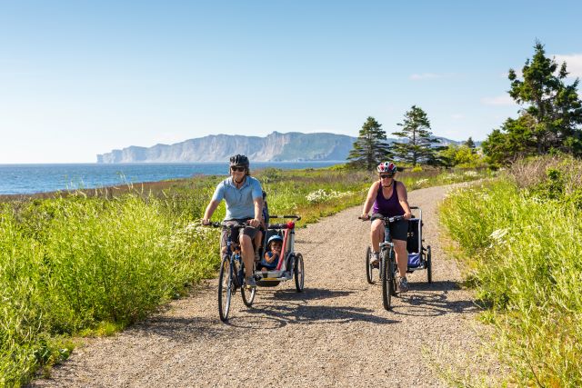 Deux cyclistes tirant des chariots pour enfants roulent sur un sentier de gravier avec les falaises au loin. 