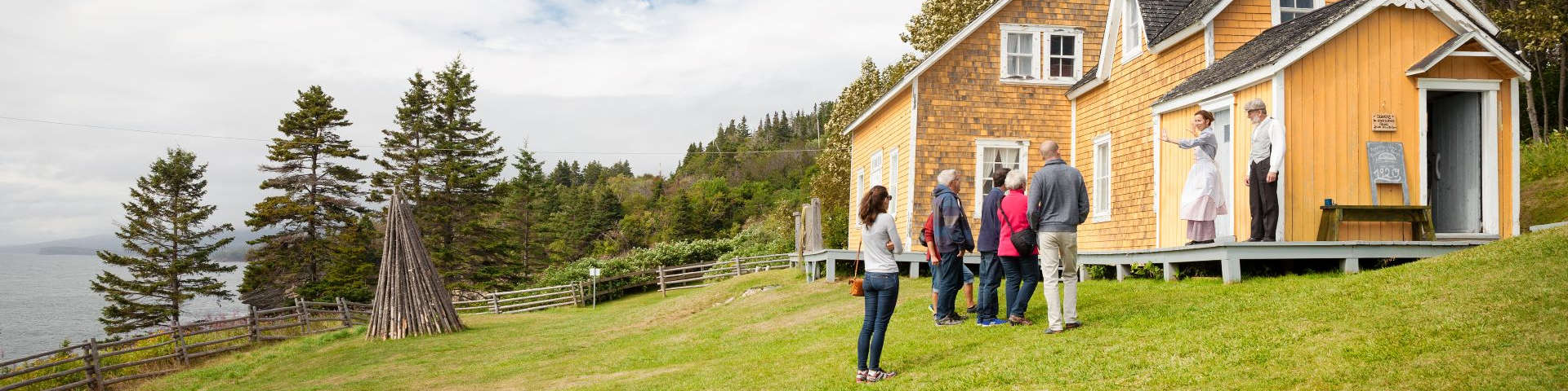 A group of visitors listen to two performers on the porch of a yellow house.