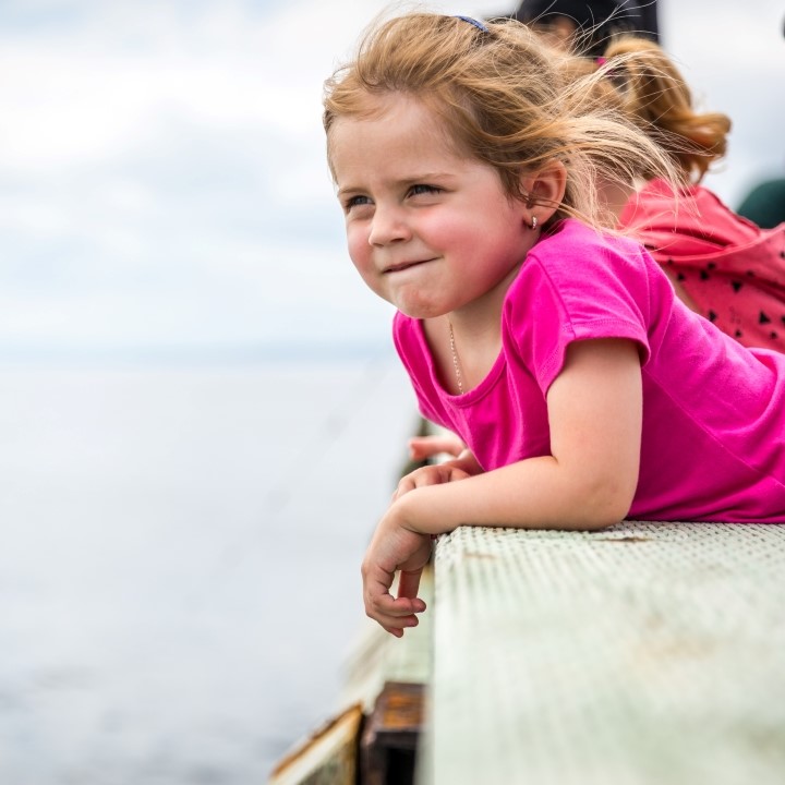 A little girl leaning against the rail of a dock looks out to the sea.