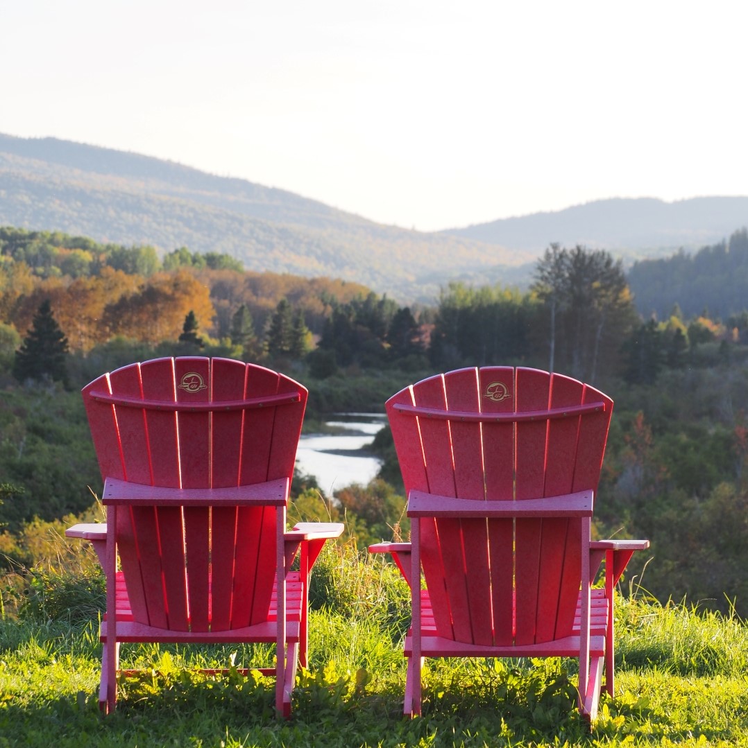 Two red Adirondack chairs in front of a valley landscape in autumn.