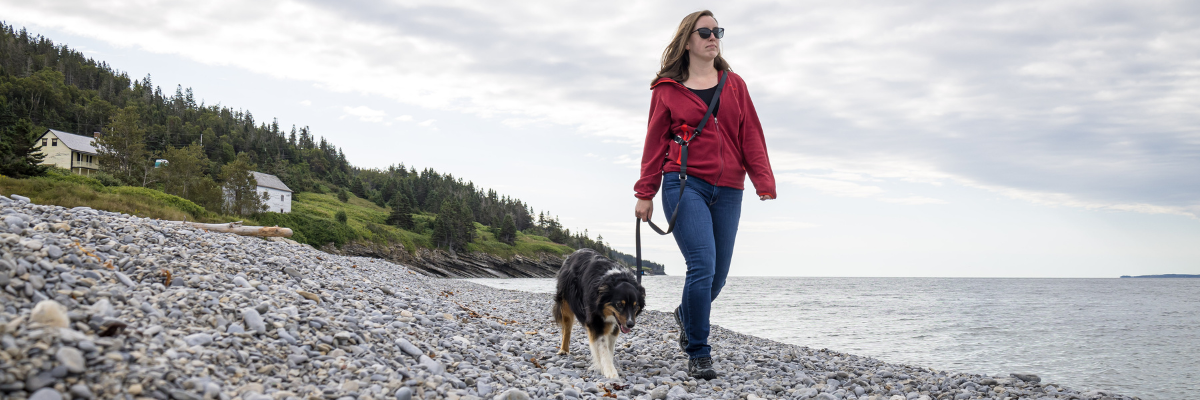 A woman walks on a pebble beach with her dog on a leash.
