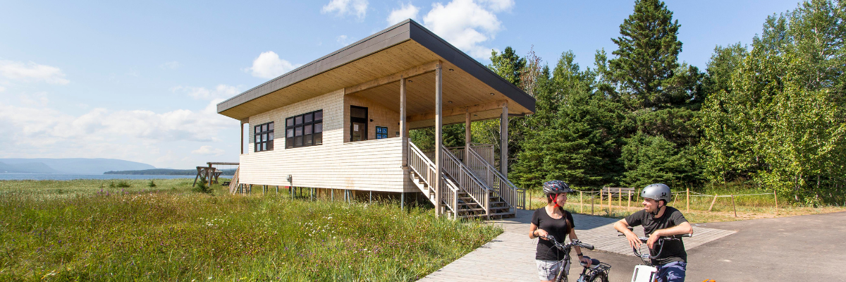 Two cyclists take a break in front of a wooden building near the beach.