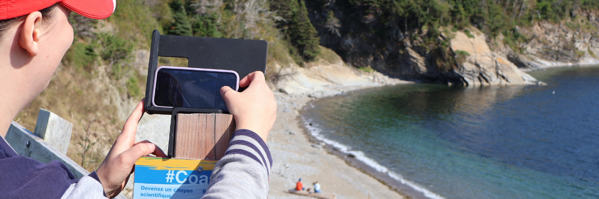 A woman places her phone on a metal stand to take a photo of the beach.