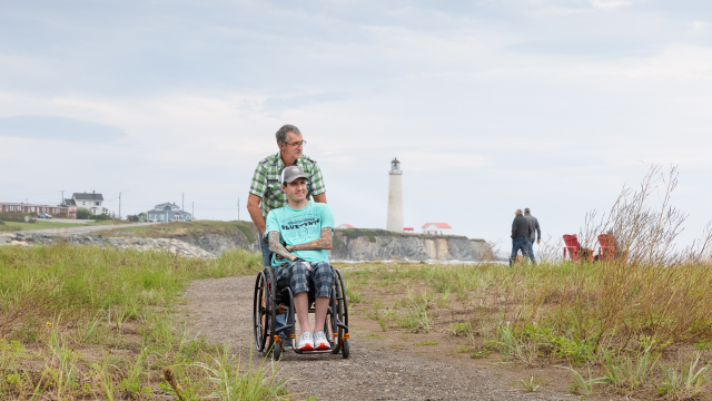 A man is pushing a young man in a wheelchair along a path by the sea.