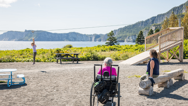 A child is playing in a playground accompanied by a lady in a wheelchair and another sitting on a bench. 