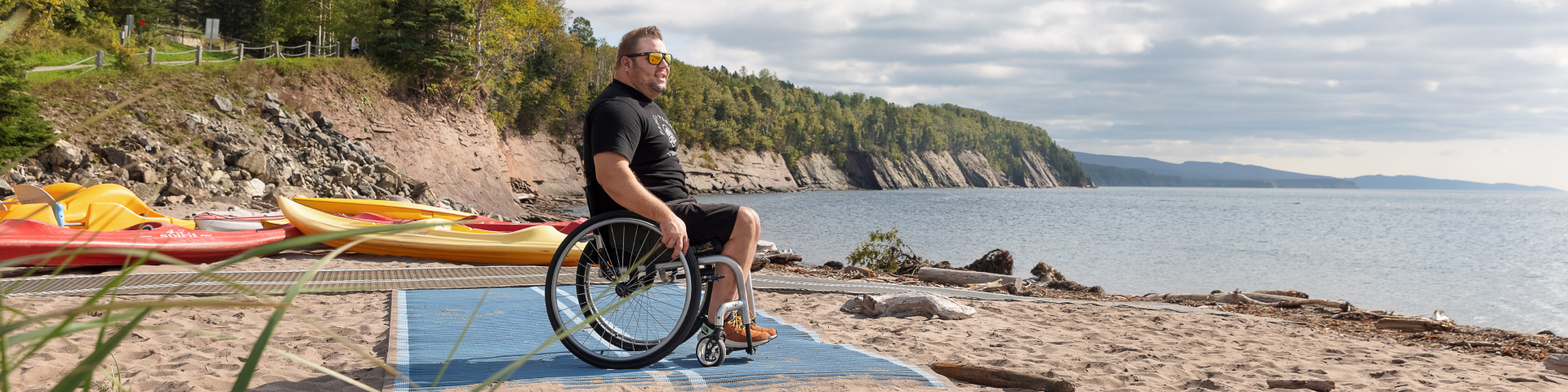 Une personne en fauteuil roulant sur un tapis d'accès de plage regarde la mer. 