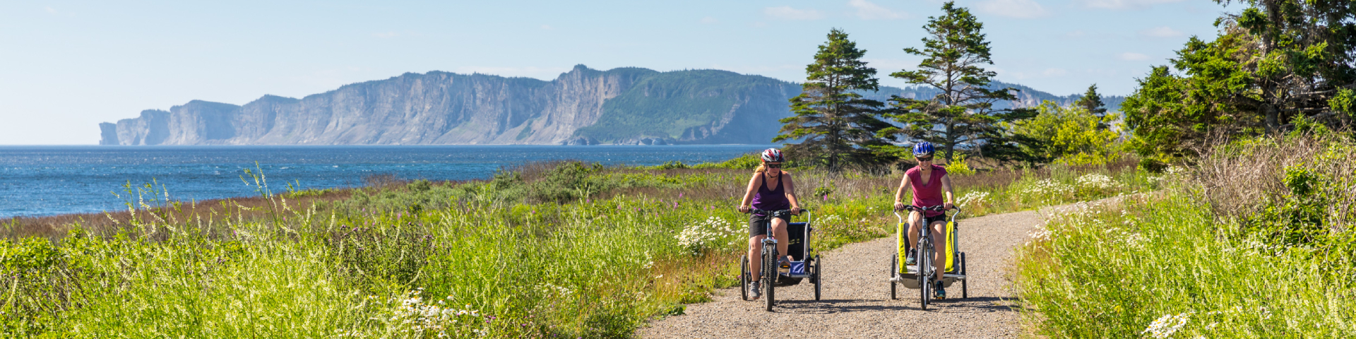 Deux cyclistes avec des poussettes pour vélo roulent sur un sentier de gravier avec les falaises en arrière-plan. 