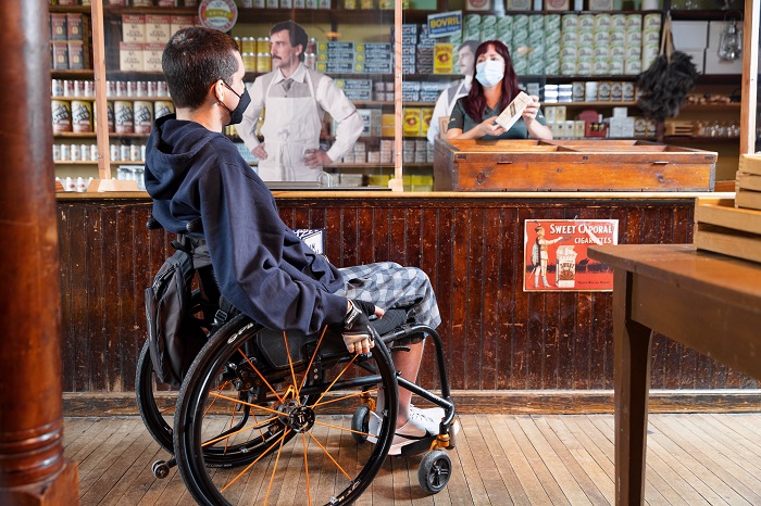 Un homme en fauteuil roulant se fait accueillir par une employée de Parcs Canada, à l'intérieur du magasin général Hyman and Sons.