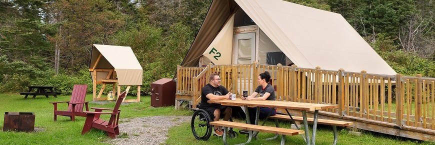 A man in a wheelchair and a woman are seated at the picnic table adapted for people with reduced mobility, in front of the oTENTik F2 tent at Petit-Gaspé campground. A ramp provides access to the tent.