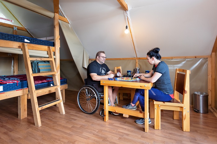 A man in a wheelchair and a woman are seated at a table inside an oTENTik tent.