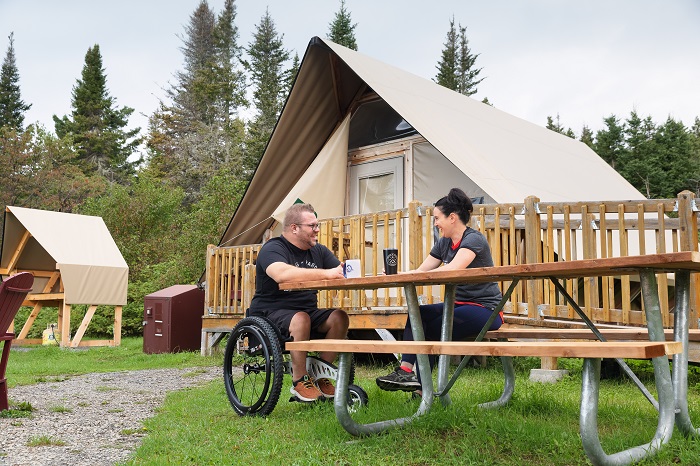 A man in a wheelchair and a woman are seated at a picnic table adapted for people with reduced mobility, in front of an oTENTik tent. 