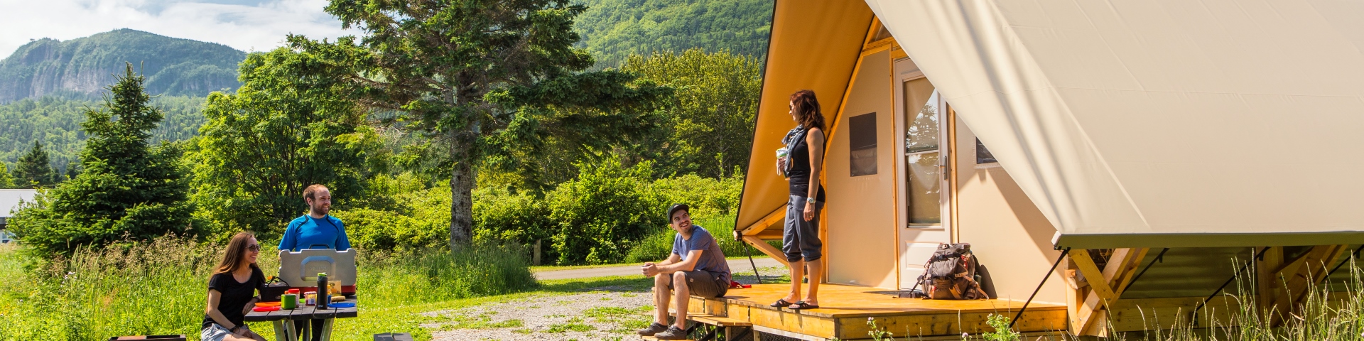 Quatre personnes, dehors, sur le site semi-boisé d'une tente oTENTik discutent. 