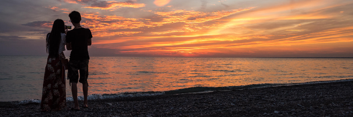 Two persons on a beach take picture of a sunset.