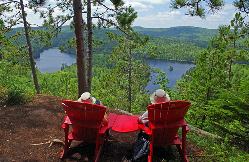 Two hikers are sitting on red chairs in front of a lake and a forest.