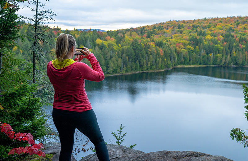 A hiker takes a picture of a lake and a forest with her cellphone.