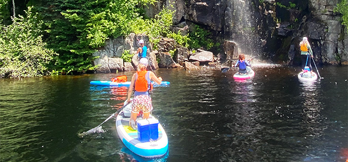 Photo provenant de l’édition 2024 de l’activité d’introduction au SUP camping. On y voit 5 personnes sur des planches à pagaie sur un lac, près d’une falaise où on voit une petite chute d’eau tomber. Autour de la chute, on y voit plusieurs conifères et feuillus verts descendant jusqu’au lac. Le tout est sous un ciel bleu.