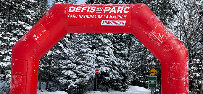 Arche rouge gonflable des Défis du Parc Nordique avec deux pistes de ski de fond classique passant en dessous. En arrière-plan, on retrouve une forêt traversée par un sentier avec des conifères enneigés à ses côtés. 