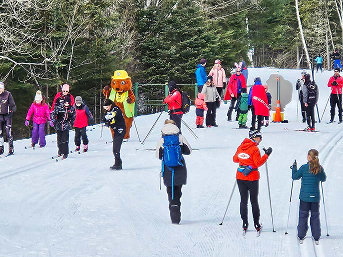 Plusieurs jeunes accompagnés par des superviseurs font du ski de fond au début du sentier #6 du parc national de la Mauricie. La mascotte Parka accompagne le groupe durant cette journée. On y voit le sentier se perdre dans la forêt en arrière-plan.