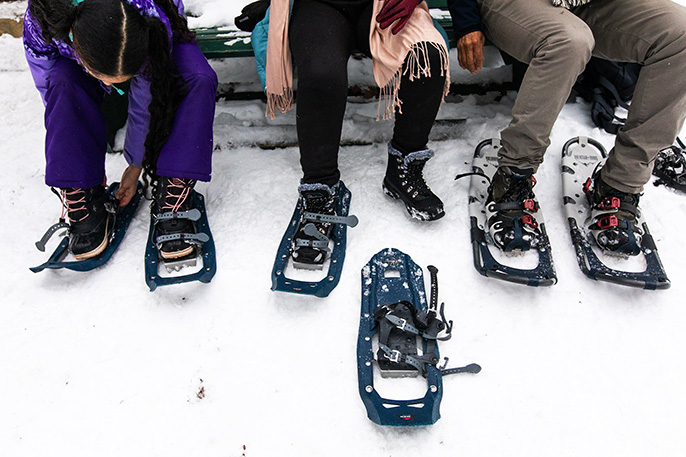 Photo d’une famille se préparant à aller en randonnée hivernale en attachant leurs raquettes sur un banc. Une jeune fille (à gauche) est dans un manteau violet et elle attache une raquette sur sa botte droite. La mère est au centre et on ne voit que ses jambes. Elle a une raquette attachée sur sa botte droite. Le père (à gauche) est assis et on ne voit que ses jambes aussi. Il a déjà ses raquettes à ses pieds. Ils sont tous assis sur un banc vert.