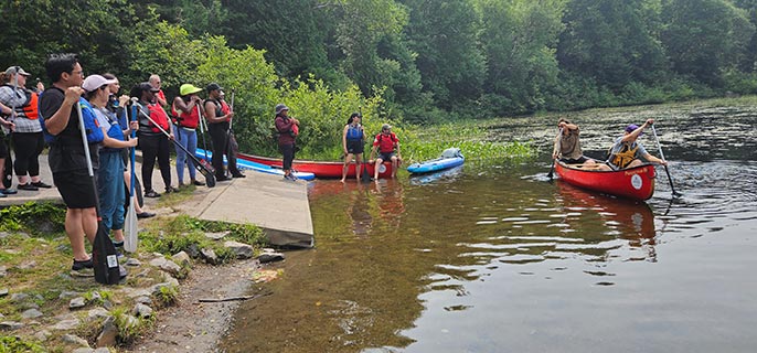 Des personnes sur la berge regardent des gens qui pagayent dans un canot sur un lac.