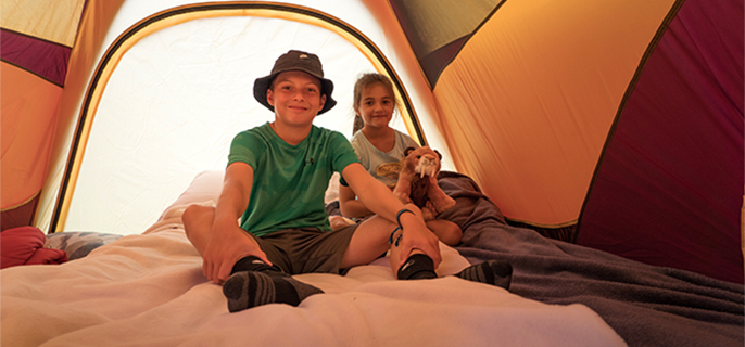 Two young children in a tent. The boy is wearing a green sweater, a black hat and brown shorts. Behind him, a little girl wears a blue sweater and holds her lion pooch.