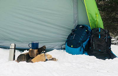 Des sacs à dos, des bûches de bois, une hache, une tasse et un thermos sont posés dans la neige devant une tente.