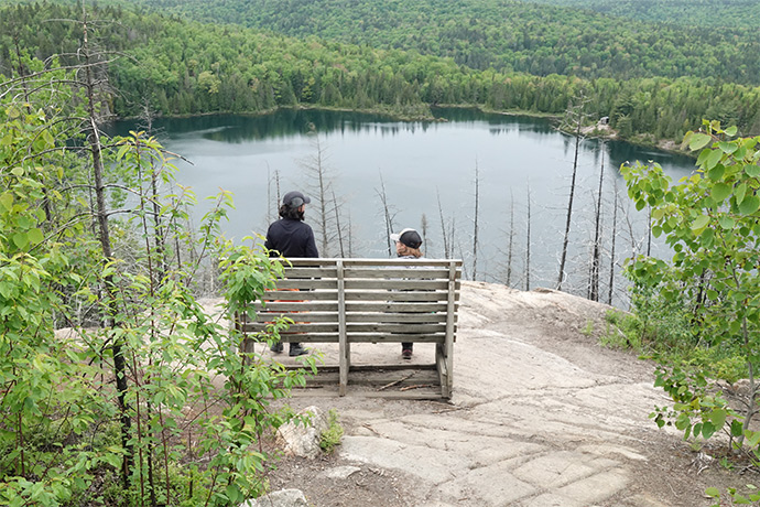 Two hikers are sitting on a bench in front of Solitaire Lake and a forest.