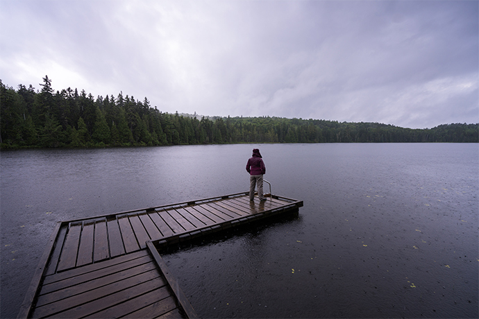 A person on a dock looks out over a lake and forest in the rain.