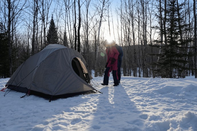 A person in a snowsuit is standing near a grey tent set up on snowy ground, in the middle of a forest of bare trees with the sun piercing through the branches in the background.